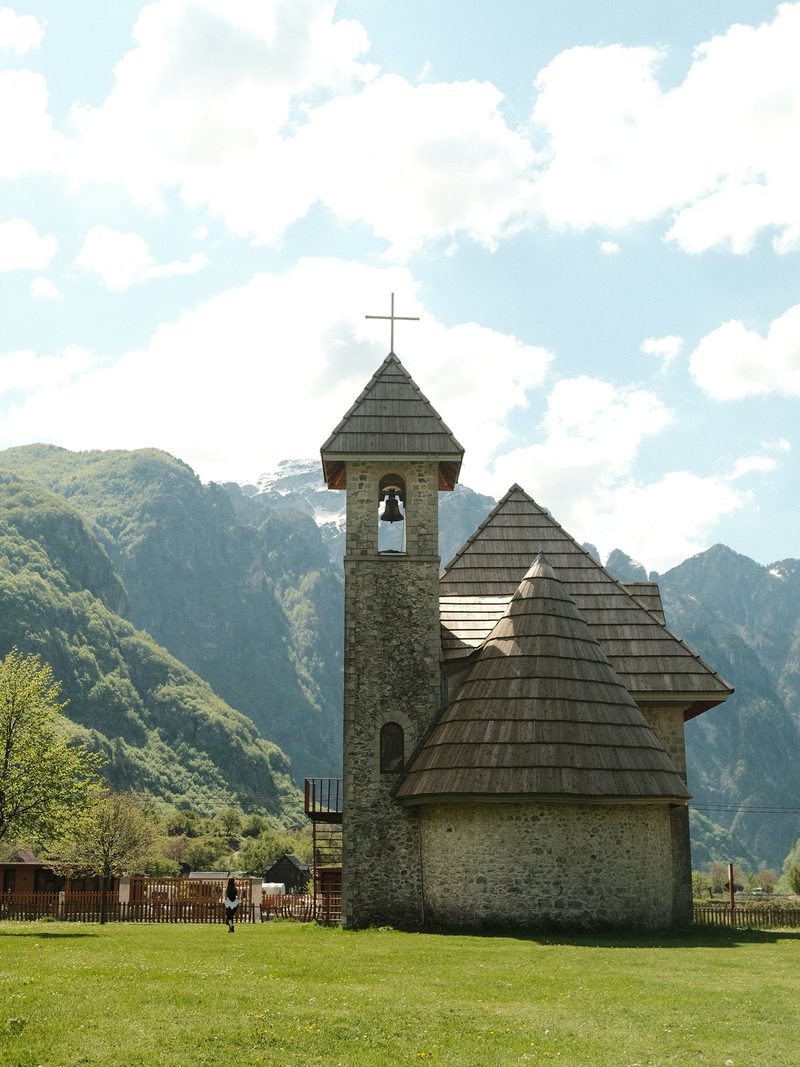 A stone church with a tall bell tower and cross stands in front of a mountain range under a blue sky with white clouds.