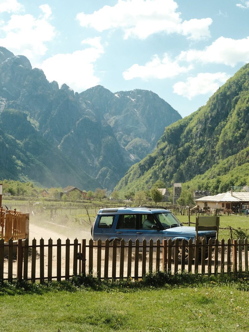 A blue SUV is parked in front of a wooden fence in a mountainous area.