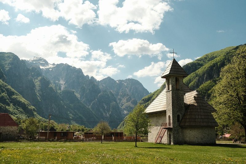 A stone church with a tall tower and cross in a mountainous valley with a grassy field in the foreground.