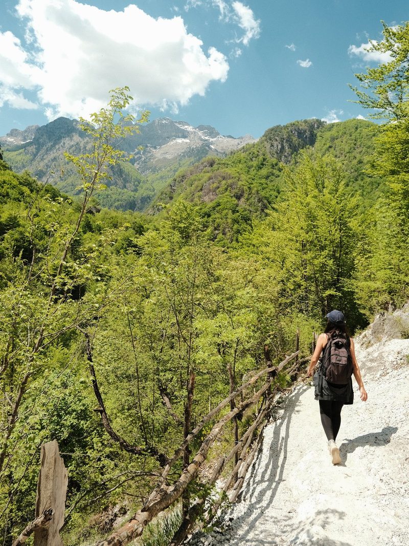 A woman hiking on a trail in a mountainous area with a fence and trees on either side.