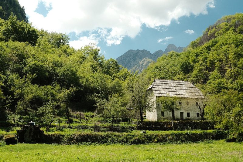 A serene rural landscape with a stone house and lush greenery, set against a backdrop of mountains and a blue sky with clouds.