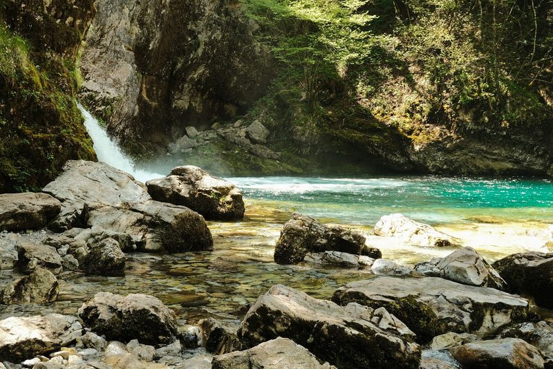 A serene waterfall cascades into a tranquil turquoise river, surrounded by lush greenery and large rocks.