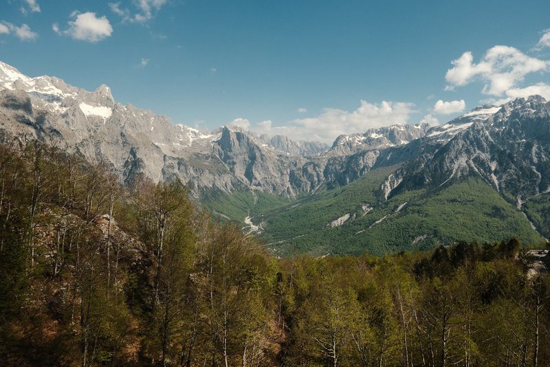A mountain range with snow-capped peaks and a valley with trees and a road.