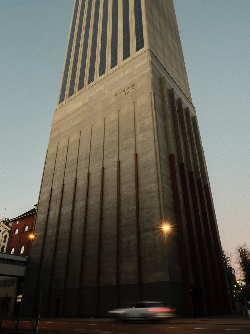 A tall, concrete building with a distinctive design, featuring a prominent vertical structure and a car driving in front of it.