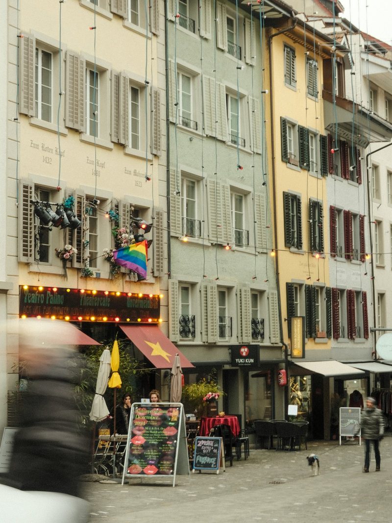 A street scene with a row of buildings, featuring a rainbow flag, a restaurant with outdoor seating, and a person walking a dog.