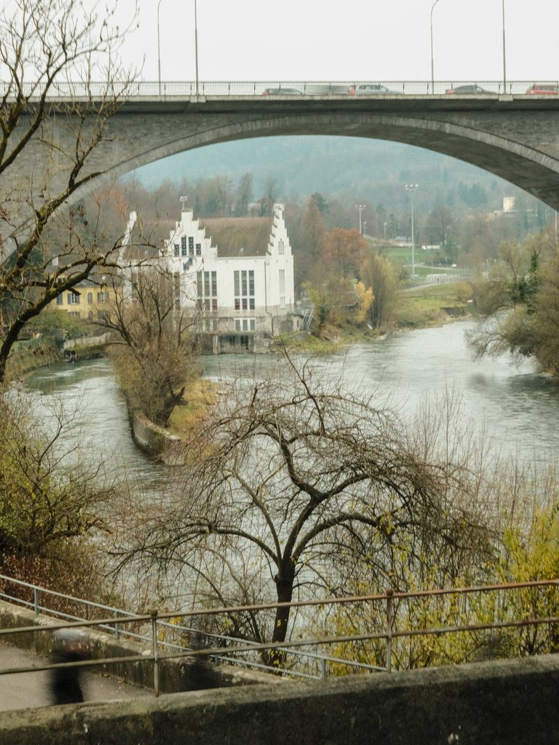 A large stone bridge spans a river, with a white building and trees in the foreground.