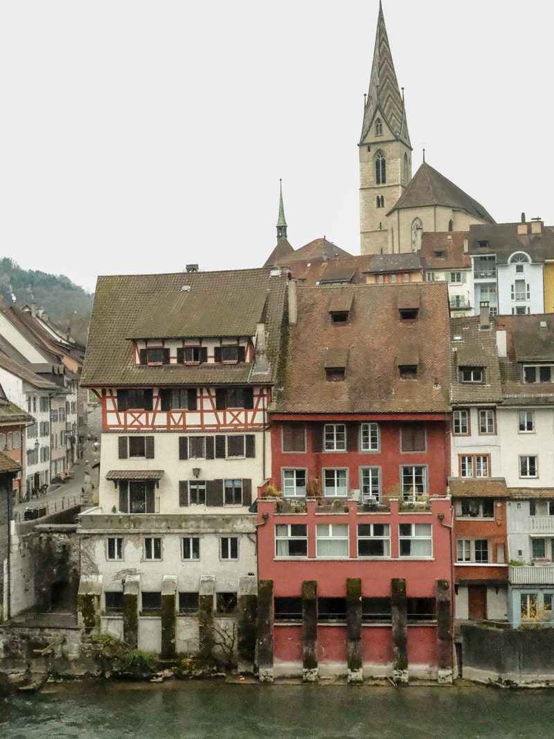 A cluster of multi-story buildings with varying architectural styles, including steeply pitched roofs and ornate facades, line a riverbank, with a tall church steeple rising above the rooftops.