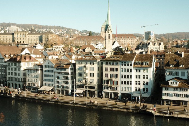 A cityscape with a river running through it, featuring a mix of old and new buildings, including a church steeple and a bridge.