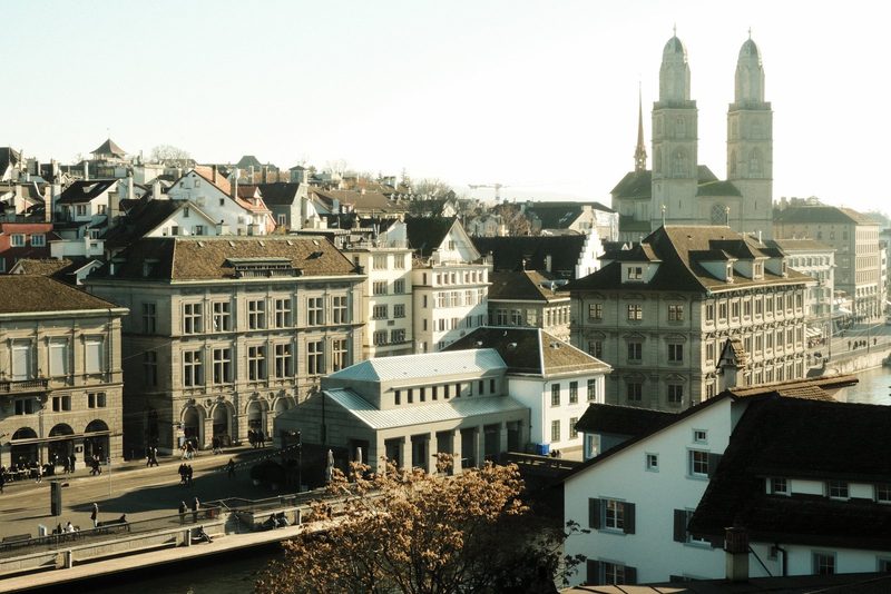 A cityscape featuring a mix of old and new buildings, with a prominent church steeple rising above the rooftops.