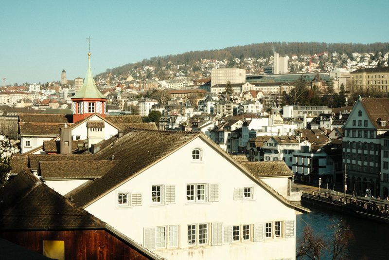 A cityscape with a mix of old and new buildings, featuring a prominent white building with a brown roof and a bridge over a body of water.