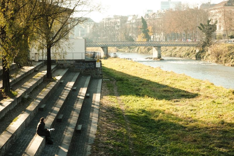 A person sits on a stone bench, holding a book, with a river and bridge in the background.