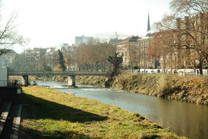 A bridge spans a river, with a grassy bank on one side and a cityscape on the other.