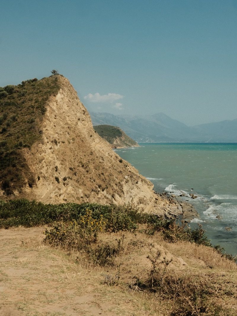 A rugged cliffside overlooks a body of water, with a mountain range in the distance.