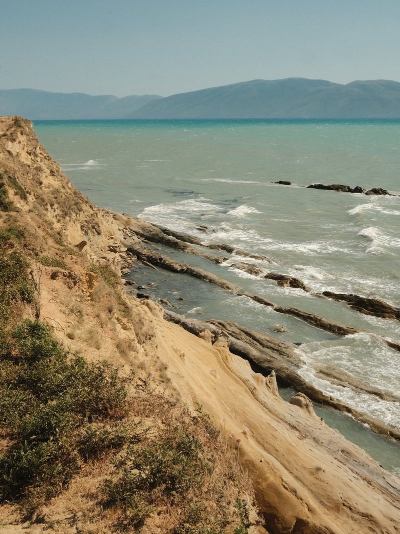 A rugged coastline with a steep cliff, featuring a sandy slope and a body of water with waves and rocks.