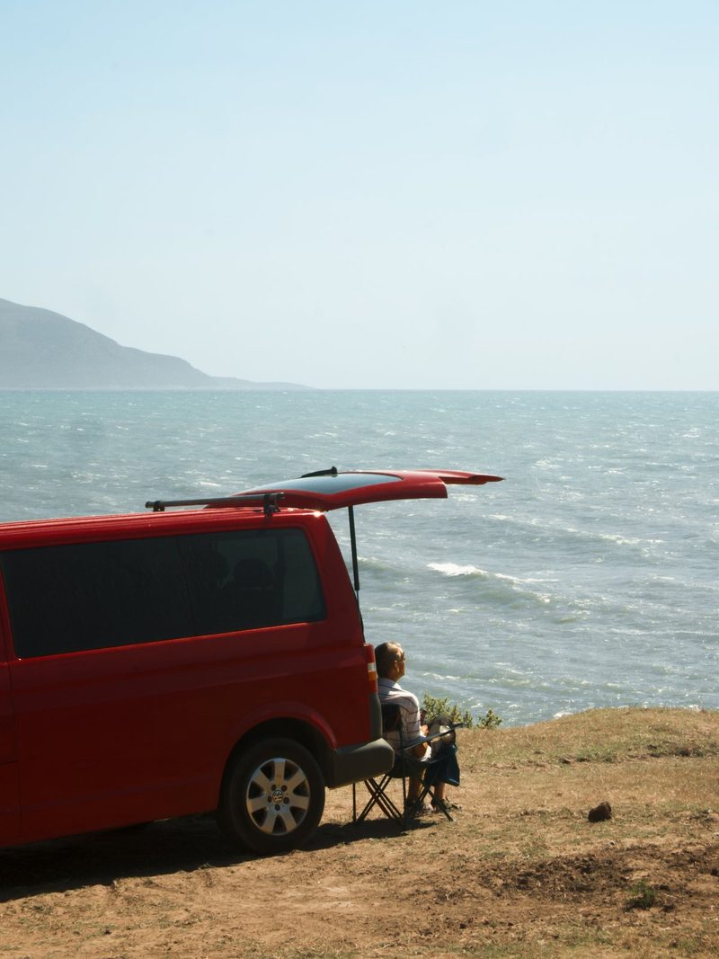 A man sits in a folding chair next to a red van with a surfboard on its roof, overlooking a body of water.