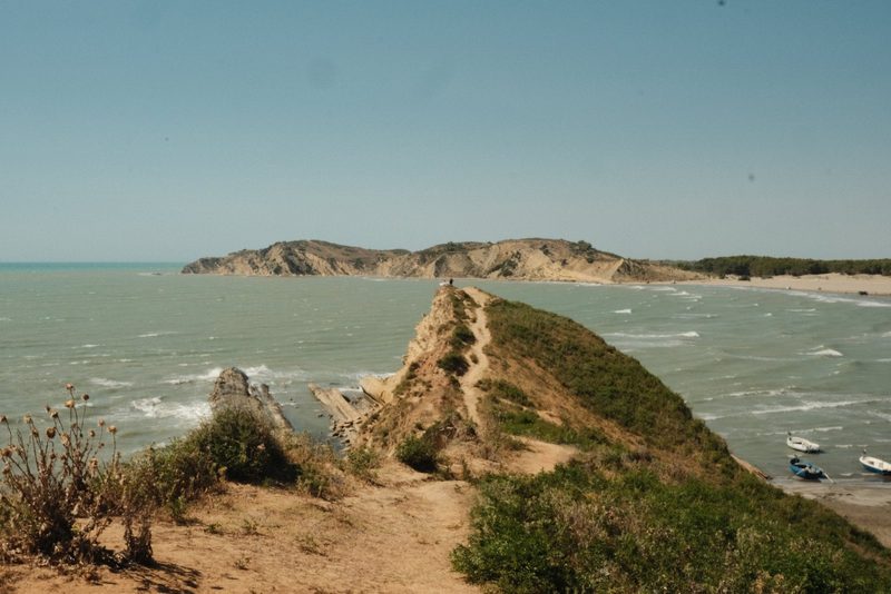A rugged coastline with a prominent cliff, a sandy beach, and a few small boats in the water.