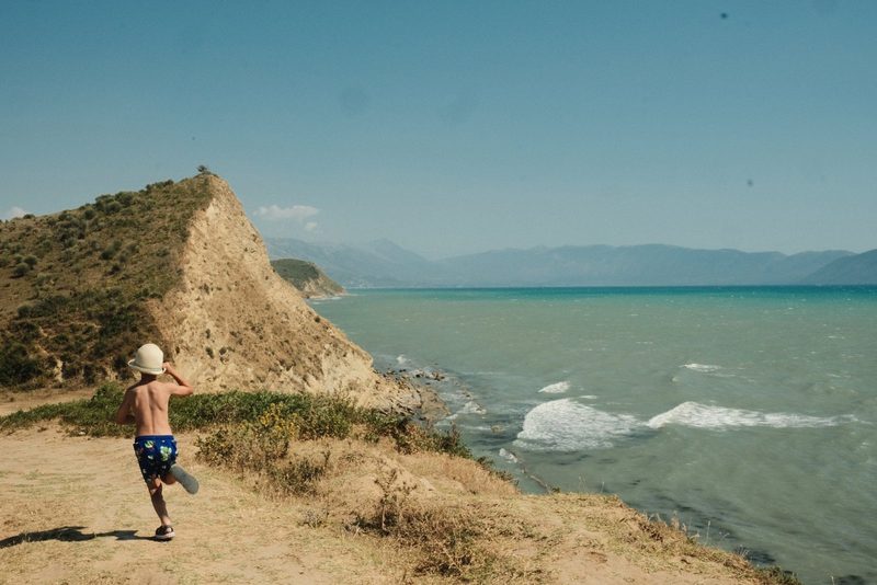 A young boy, shirtless and wearing a hat, blue shorts, and sandals, runs along a dirt path near a body of water, with a rocky cliff and hills in the background.