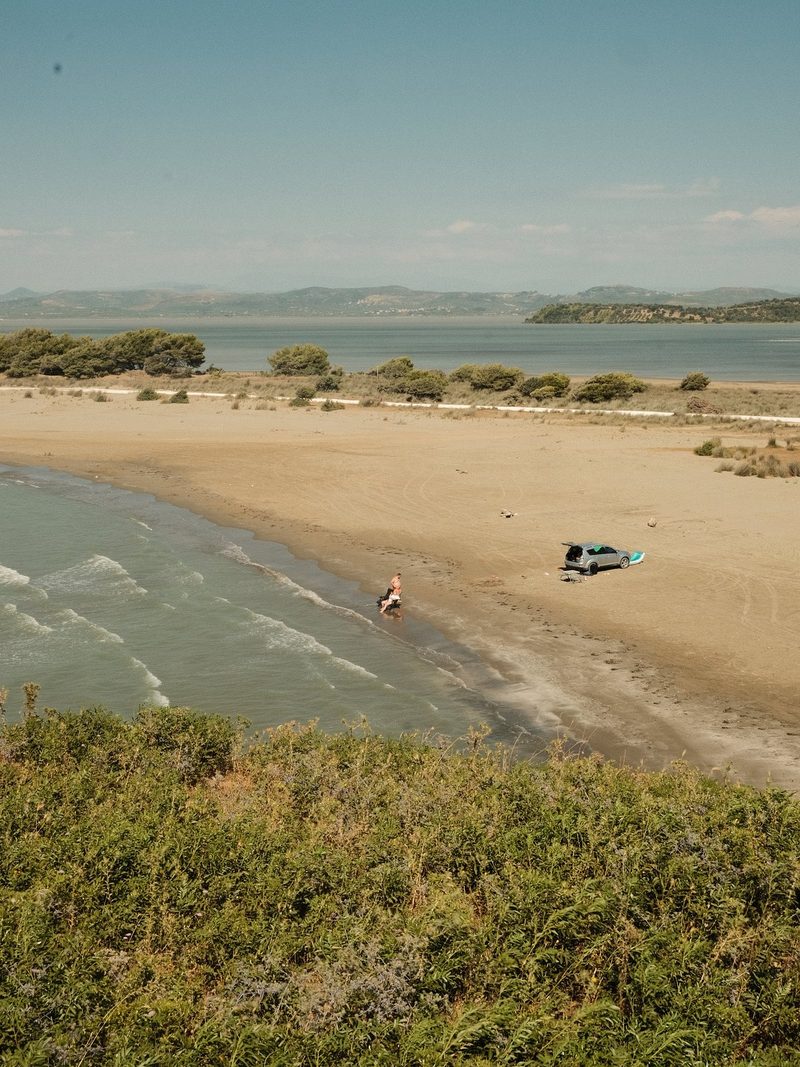 A woman in a swimsuit sits on a beach, with a car parked nearby.
