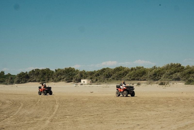 Two people ride orange all-terrain vehicles on a sandy terrain, with a line of trees in the background.