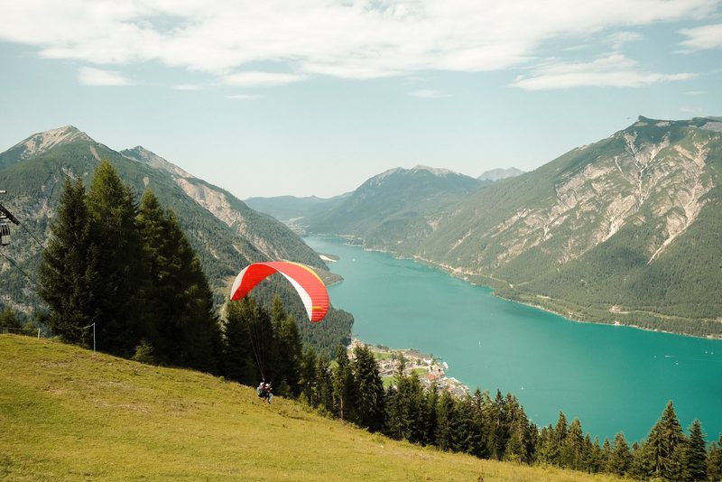 A person in a red and white parachute is descending towards a body of water, surrounded by mountains and trees.