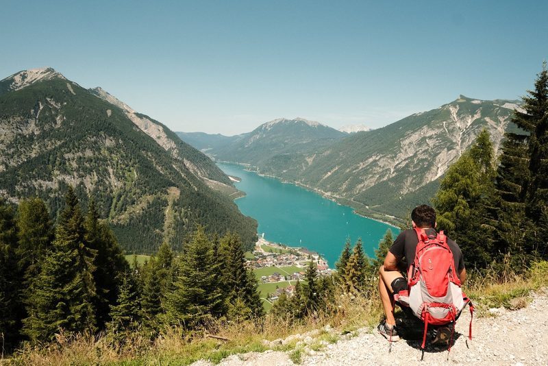 A man sits on a rocky outcropping, gazing out at a serene lake nestled among majestic mountains and lush evergreen trees.