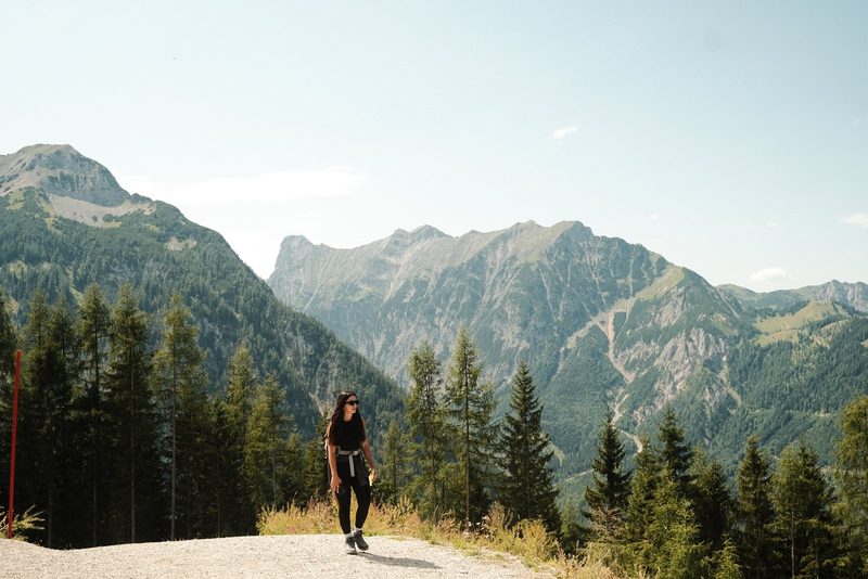 A woman walks along a gravel path, surrounded by trees and mountains in the background.