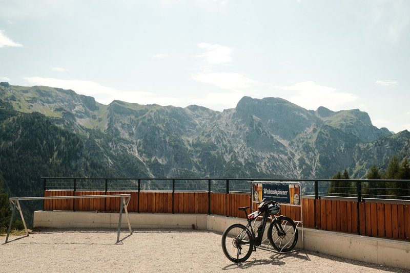 A mountain range with a prominent peak, set against a backdrop of a blue sky with scattered clouds. In the foreground, a wooden fence and a bicycle are visible.