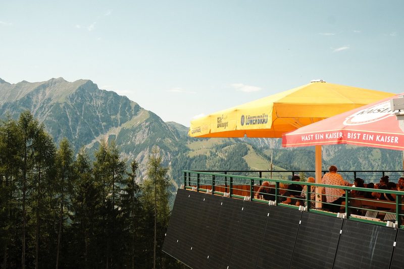 A group of people sit on a balcony with a railing, overlooking a mountain range and a yellow and red umbrella.