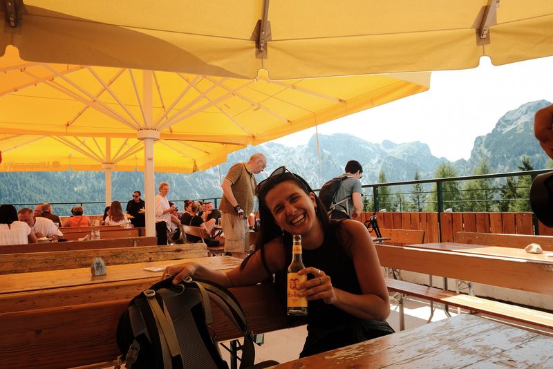A woman sits at a wooden table, holding a bottle of beer and wearing a black tank top, with a backpack on the table in front of her.
