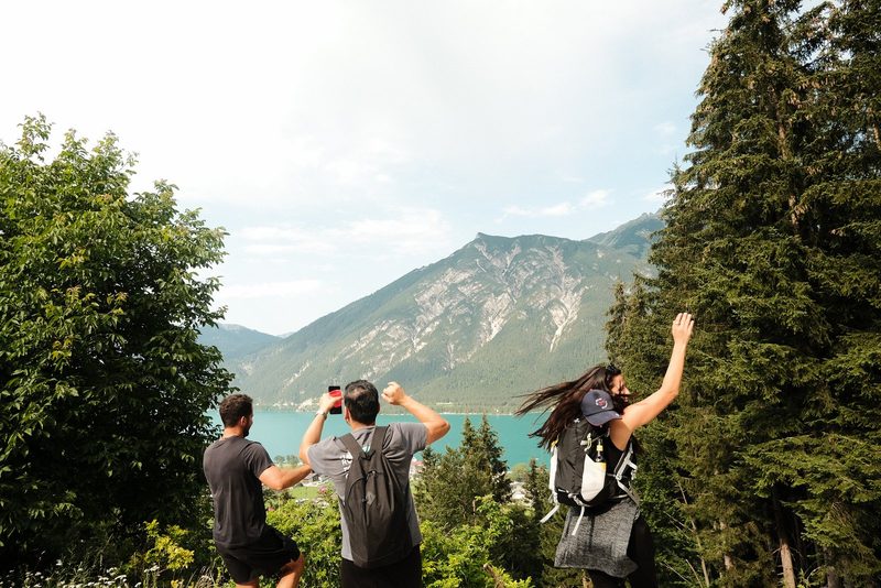 Three people stand in front of a mountainous landscape, with two of them holding drinks and one jumping in the air.