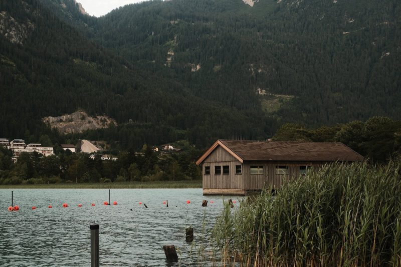 A wooden building with a sloping roof and a small windowed section, partially submerged in a body of water, with a grassy bank and a mountainous backdrop.