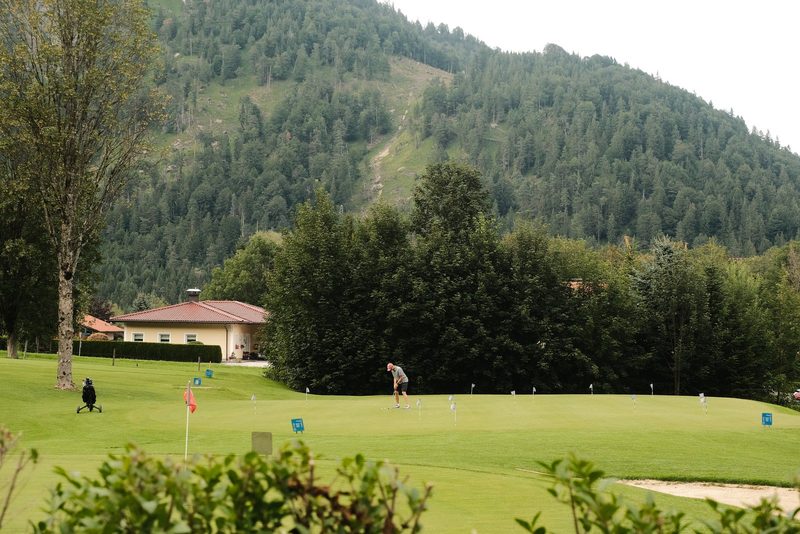 A man is putting on a golf course, with a house and trees in the background.